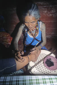 The Last Kalinga tattooist Whang Od hand-tapping a new design on Elaine Tima in a dark back room at the Batok Festival. She uses the rear-end of her kisi or tattooing tool to stencil the design. The kisi consists of a piece of carabao (water buffalo) horn bent by fire resembling the letter L holding a row of four or five razor-sharp thorns or needles in its head. Elaine's father is a noted Kalinga anthropologist. At other times, Whang Od uses an orange thorn attached to a small bamboo stick to make her tattoos.